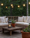 A cozy farmhouse deck features deep-seated wicker furniture with cream cushions and plaid throw pillows, illuminated by warm string lights and a metal lantern. Terracotta pots with lush greenery and a rustic wooden coffee table complete the inviting outdoor scene. — alternate angle