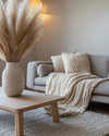 A cozy modern landscape living room featuring a warm gray sofa, a chunky cream knit throw, a light oak coffee table, and a large ceramic vase with dried pampas grass, all illuminated by soft natural light.