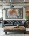 An industrial landscape living room featuring a charcoal distressed leather Chesterfield sofa, a reclaimed wood coffee table on caster wheels, and a large abstract painting in grey and burnt sienna tones, under exposed metal pendant lighting. — alternate angle