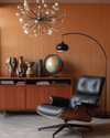 A sophisticated Mid-Century Modern library featuring an Eames Lounge Chair in black leather and walnut, an Arco floor lamp arching over, and a teak credenza displaying abstract ceramic vases and a vintage globe, all under the warm glow of a Sputnik chandelier. — styling and décor detail