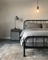 An industrial bedroom featuring a black metal bed frame, a grey washed linen duvet, and an exposed Edison bulb pendant light hanging from the ceiling. A dark wood nightstand holds a concrete-look lamp, and a large, distressed area rug covers the light grey floor.