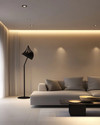 A modern living room at dusk with layered lighting, featuring recessed ceiling lights, a sleek matte black floor lamp beside a light gray sofa, and a sculptural pendant light illuminating a minimalist coffee table. Warm white light creates an inviting ambiance. — styling and décor detail