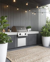 A modern outdoor kitchen setup featuring a charcoal gray modular grill station with a light concrete-effect countertop, brushed stainless steel accents, and black string lights strung overhead. Large white ceramic planters with architectural green plants frame the area, complemented by a woven outdoor rug in a subtle geometric pattern.