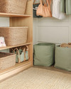 Boho closet storage with open mango wood shelving, large woven rattan baskets for toys, and canvas bins in muted sage green. A low-pile jute rug sits on the floor, and a small, dedicated hook is placed at a child's height.