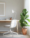 A bright Scandinavian home office featuring a light oak desk, a white ergonomic chair, and a minimalist white floor lamp. A large fiddle leaf fig plant in a terracotta pot sits beside the desk, complementing the warm grey rug and muted artwork leaning against the wall. — alternate angle