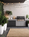A modern outdoor kitchen setup featuring a charcoal gray modular grill station with a light concrete-effect countertop, brushed stainless steel accents, and black string lights strung overhead. Large white ceramic planters with architectural green plants frame the area, complemented by a woven outdoor rug in a subtle geometric pattern. — close-up of textures and materials