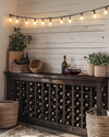 A cozy farmhouse wine cellar featuring a dark stained wooden wine rack filled with bottles, a rustic console table with a decanter, and warm string lights with Edison bulbs draped across a temporary shiplap wall in antique white. A few woven baskets and ceramic planters with subtle greenery complete the inviting, rustic aesthetic. — styling and décor detail