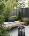 A Scandinavian backyard featuring a light grey wooden deck, a simple slatted teak bench with a charcoal grey outdoor cushion, and a minimalist black metal lantern. Greenery includes potted evergreen shrubs and ornamental grasses in terracotta pots, creating a serene, natural ambiance. — styling and décor detail