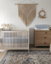 Coastal baby and kids' room with a white oak crib, a large seagrass rug, a rattan changing table, and a macrame wall hanging above a dresser. Soft white walls are accented with muted blue textiles and natural wood elements. — styling and décor detail