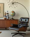 A sophisticated Mid-Century Modern library featuring an Eames Lounge Chair in black leather and walnut, an Arco floor lamp arching over, and a teak credenza displaying abstract ceramic vases and a vintage globe, all under the warm glow of a Sputnik chandelier. — alternate angle