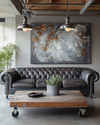 An industrial landscape living room featuring a charcoal distressed leather Chesterfield sofa, a reclaimed wood coffee table on caster wheels, and a large abstract painting in grey and burnt sienna tones, under exposed metal pendant lighting. — styling and décor detail