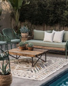 A boho poolside area featuring a mix of vintage wrought-iron chairs in sage green and a modern low-slung outdoor sofa with natural linen cushions. A reclaimed wooden coffee table sits on a layered outdoor rug with geometric patterns, surrounded by potted succulents and woven rattan accents.