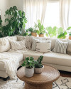 A cozy boho family room featuring a cream sectional sofa adorned with macrame and striped throw pillows, a round rattan coffee table, and an abundance of lush green plants in woven and ceramic planters, all bathed in natural light from a large window with sheer linen curtains. — close-up of textures and materials