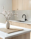 A serene Japandi kitchen featuring light ash wood cabinetry, a white quartz countertop, and minimalist black metal fixtures. A single ceramic vase holds delicate cherry blossom branches, and a natural woven placemat sits on the island. — styling and décor detail