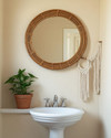 A cozy boho powder room featuring a round rattan mirror above a white pedestal sink, a small Pothos plant in a terracotta pot, and a macrame wall hanging on a light beige wall. — alternate angle