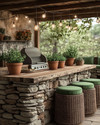 A cozy rustic outdoor kitchen featuring a stacked stone island with a natural wood countertop, a built-in grill, and string lights draped overhead. Terracotta pots with herbs sit on the counter, and comfortable wicker stools with green cushions are pulled up to the bar. — styling and décor detail