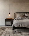 An industrial bedroom featuring a black metal bed frame, a grey washed linen duvet, and an exposed Edison bulb pendant light hanging from the ceiling. A dark wood nightstand holds a concrete-look lamp, and a large, distressed area rug covers the light grey floor. — close-up of textures and materials
