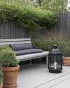 A Scandinavian backyard featuring a light grey wooden deck, a simple slatted teak bench with a charcoal grey outdoor cushion, and a minimalist black metal lantern. Greenery includes potted evergreen shrubs and ornamental grasses in terracotta pots, creating a serene, natural ambiance. — alternate angle