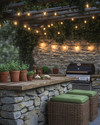 A cozy rustic outdoor kitchen featuring a stacked stone island with a natural wood countertop, a built-in grill, and string lights draped overhead. Terracotta pots with herbs sit on the counter, and comfortable wicker stools with green cushions are pulled up to the bar. — close-up of textures and materials