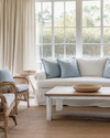 A serene coastal sunroom featuring a slipcovered sofa in a creamy off-white, paired with rattan chairs and a bleached oak coffee table. Soft dusty blue throw pillows and a woven jute rug add subtle color accents, all illuminated by ample natural light filtering through sheer linen curtains. — alternate angle