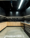A clean industrial garage featuring polished concrete floors, birch plywood cabinets, and black perforated steel wall panels for tool organization. Overhead LED strip lights illuminate a stainless steel workbench, with a few carefully arranged silver and black tools. — styling and décor detail