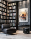 A modern library featuring floor-to-ceiling matte black bookshelves filled with books, a low-profile charcoal leather armchair, a black marble side table, and a large abstract art piece on a wall painted in Benjamin Moore's Gray Owl, with warm task lighting. — styling and décor detail