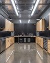 A clean industrial garage featuring polished concrete floors, birch plywood cabinets, and black perforated steel wall panels for tool organization. Overhead LED strip lights illuminate a stainless steel workbench, with a few carefully arranged silver and black tools. — alternate angle