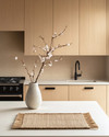 A serene Japandi kitchen featuring light ash wood cabinetry, a white quartz countertop, and minimalist black metal fixtures. A single ceramic vase holds delicate cherry blossom branches, and a natural woven placemat sits on the island. — close-up of textures and materials