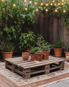 A budget-friendly Boho Backyard featuring a low wooden pallet coffee table, layered vintage rugs in muted terracotta and beige, and an assortment of potted green plants in upcycled terra cotta and ceramic containers, all illuminated by warm solar string lights strung overhead. — styling and décor detail