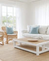 A serene coastal sunroom featuring a slipcovered sofa in a creamy off-white, paired with rattan chairs and a bleached oak coffee table. Soft dusty blue throw pillows and a woven jute rug add subtle color accents, all illuminated by ample natural light filtering through sheer linen curtains. — close-up of textures and materials