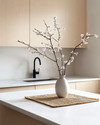 A serene Japandi kitchen featuring light ash wood cabinetry, a white quartz countertop, and minimalist black metal fixtures. A single ceramic vase holds delicate cherry blossom branches, and a natural woven placemat sits on the island. — alternate angle