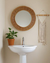 A cozy boho powder room featuring a round rattan mirror above a white pedestal sink, a small Pothos plant in a terracotta pot, and a macrame wall hanging on a light beige wall. — close-up of textures and materials