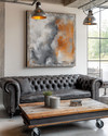 An industrial landscape living room featuring a charcoal distressed leather Chesterfield sofa, a reclaimed wood coffee table on caster wheels, and a large abstract painting in grey and burnt sienna tones, under exposed metal pendant lighting.