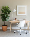 A bright Scandinavian home office featuring a light oak desk, a white ergonomic chair, and a minimalist white floor lamp. A large fiddle leaf fig plant in a terracotta pot sits beside the desk, complementing the warm grey rug and muted artwork leaning against the wall. — close-up of textures and materials