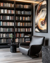 A modern library featuring floor-to-ceiling matte black bookshelves filled with books, a low-profile charcoal leather armchair, a black marble side table, and a large abstract art piece on a wall painted in Benjamin Moore's Gray Owl, with warm task lighting.
