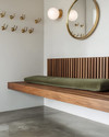 A Mid-Century Modern mudroom featuring a walnut slatted bench, polished concrete flooring, and an olive green upholstered cushion. Brass hooks line the wall above, with a circular mirror and a small white globe sconce adding modern accents.
