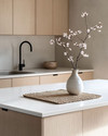 A serene Japandi kitchen featuring light ash wood cabinetry, a white quartz countertop, and minimalist black metal fixtures. A single ceramic vase holds delicate cherry blossom branches, and a natural woven placemat sits on the island.