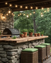 A cozy rustic outdoor kitchen featuring a stacked stone island with a natural wood countertop, a built-in grill, and string lights draped overhead. Terracotta pots with herbs sit on the counter, and comfortable wicker stools with green cushions are pulled up to the bar.