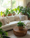 A cozy boho family room featuring a cream sectional sofa adorned with macrame and striped throw pillows, a round rattan coffee table, and an abundance of lush green plants in woven and ceramic planters, all bathed in natural light from a large window with sheer linen curtains. — styling and décor detail