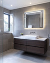 Modern bathroom with a floating dark wood vanity, white vessel sink, and large format grey wall tiles. Integrated LED mirror lighting and recessed ceiling lights illuminate the space, complemented by polished chrome fixtures. — alternate angle
