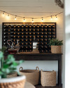 A cozy farmhouse wine cellar featuring a dark stained wooden wine rack filled with bottles, a rustic console table with a decanter, and warm string lights with Edison bulbs draped across a temporary shiplap wall in antique white. A few woven baskets and ceramic planters with subtle greenery complete the inviting, rustic aesthetic.