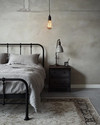 An industrial bedroom featuring a black metal bed frame, a grey washed linen duvet, and an exposed Edison bulb pendant light hanging from the ceiling. A dark wood nightstand holds a concrete-look lamp, and a large, distressed area rug covers the light grey floor. — alternate angle