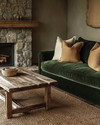 A Rustic Landscape living room featuring a deep forest green velvet sofa, a reclaimed wood coffee table, and a rough-hewn stone fireplace. Natural light illuminates the warm greige walls, complemented by woven jute rugs and subtle ochre throw pillows. — alternate angle