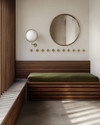 A Mid-Century Modern mudroom featuring a walnut slatted bench, polished concrete flooring, and an olive green upholstered cushion. Brass hooks line the wall above, with a circular mirror and a small white globe sconce adding modern accents. — alternate angle