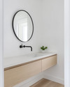 A bright Scandinavian powder room with white walls, a light wood floating vanity, a minimalist round mirror, and a single black wall-mounted faucet, accented by a small green plant. — alternate angle