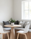 A small, bright Scandinavian breakfast nook featuring a light wood pedestal table, two minimalist white chairs, and a built-in banquette with grey linen cushions. A small potted plant sits on the table, and white walls create an airy, open feel. — close-up of textures and materials
