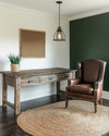 A cozy rustic home office featuring a reclaimed oak desk, a distressed cognac leather chair, and a jute rug. Walls are painted a warm off-white, complemented by a deep forest green accent wall, with an iron pendant light illuminating the space. — styling and décor detail