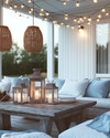 A coastal patio at dusk with warm string lights strung overhead, rattan pendant lights, and hurricane lanterns on a weathered wood table, surrounded by comfortable white cushions and soft blue throws. — styling and décor detail