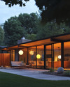 A Mid-Century Modern exterior at dusk, featuring a low-slung roofline, large glass windows, and warm glow from spherical globe sconces flanking the entry. A Sputnik chandelier is visible under a covered patio, illuminating a seating area with clean-lined furniture and a natural wood fence. — alternate angle
