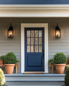 A cozy traditional exterior featuring a warm gray facade with creamy white trim, a deep blue front door, and classic black lantern sconces flanking the entrance. Potted green plants in terracotta pots sit on either side of the door, and a traditional wooden bench offers a welcoming touch. — styling and décor detail